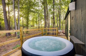 A hot tub in a wooded area with a wooden deck and a building in the background. - Cozy Onalaska Cabin ~ 2 Mi to Lake Livingston! (Onalaska)