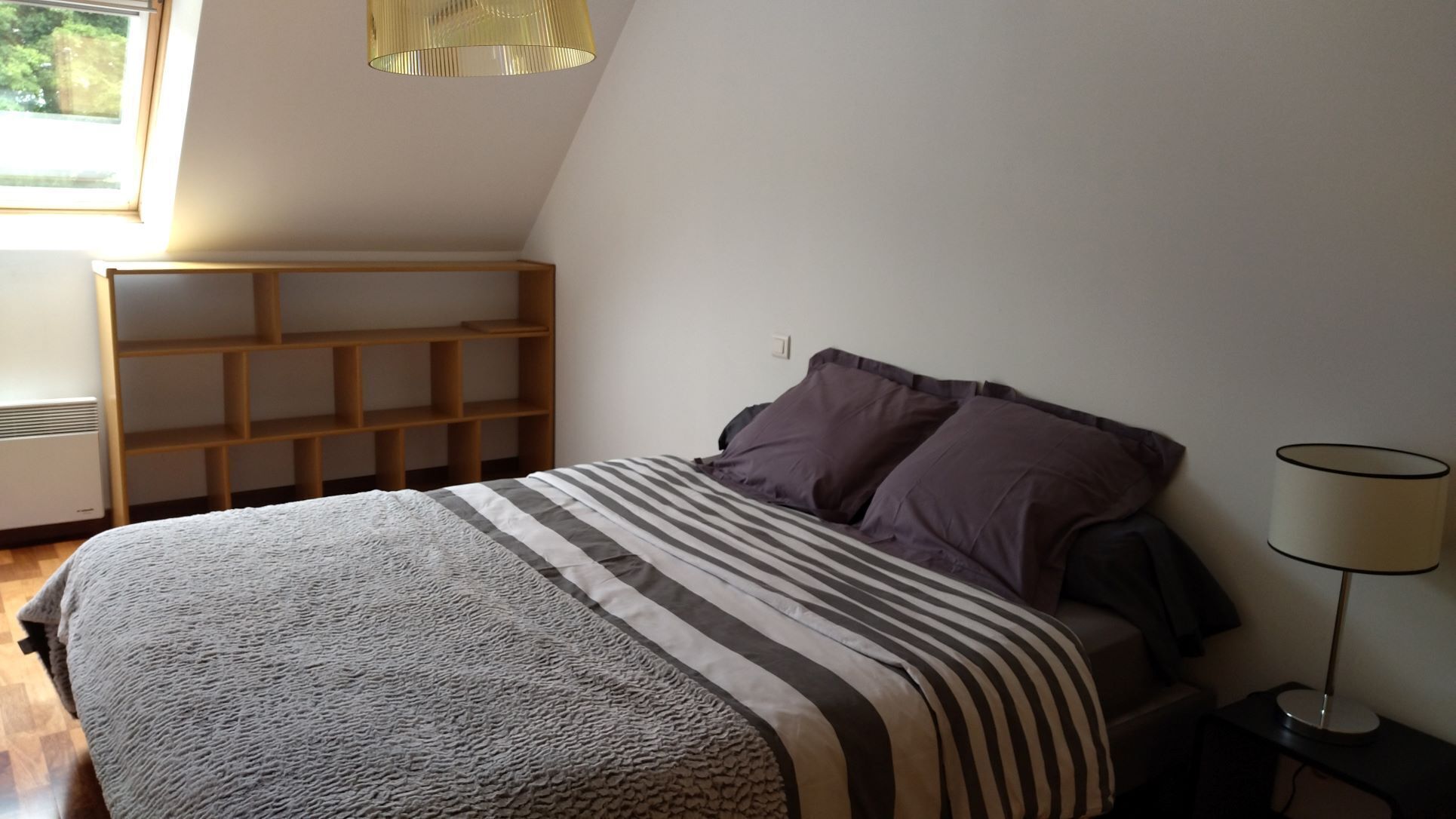 A bedroom with a bed, a wooden shelf unit, a lamp, and a window.