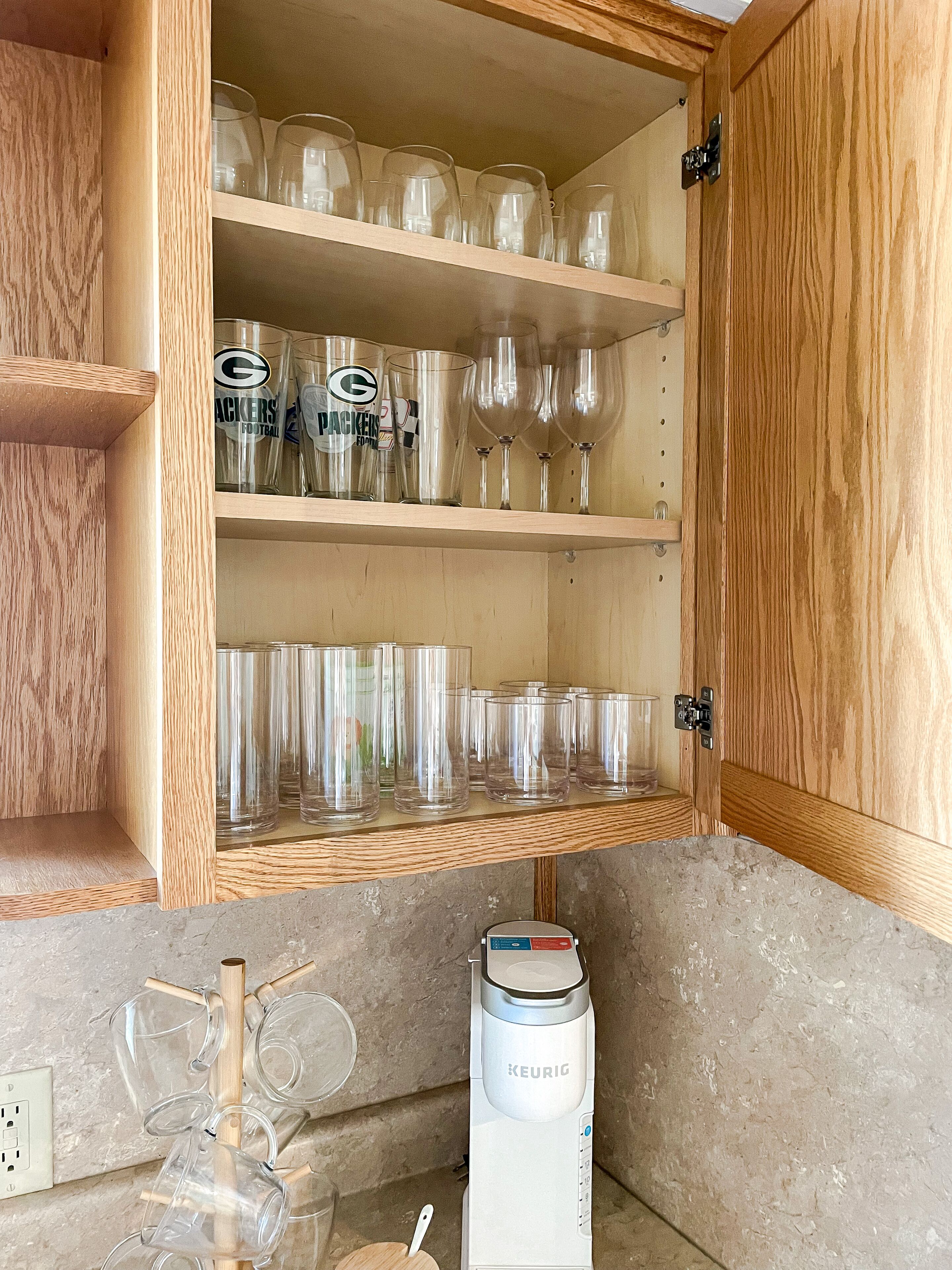 A wooden cabinet with shelves holding clear drinking glasses and a Keurig coffee maker on the countertop.