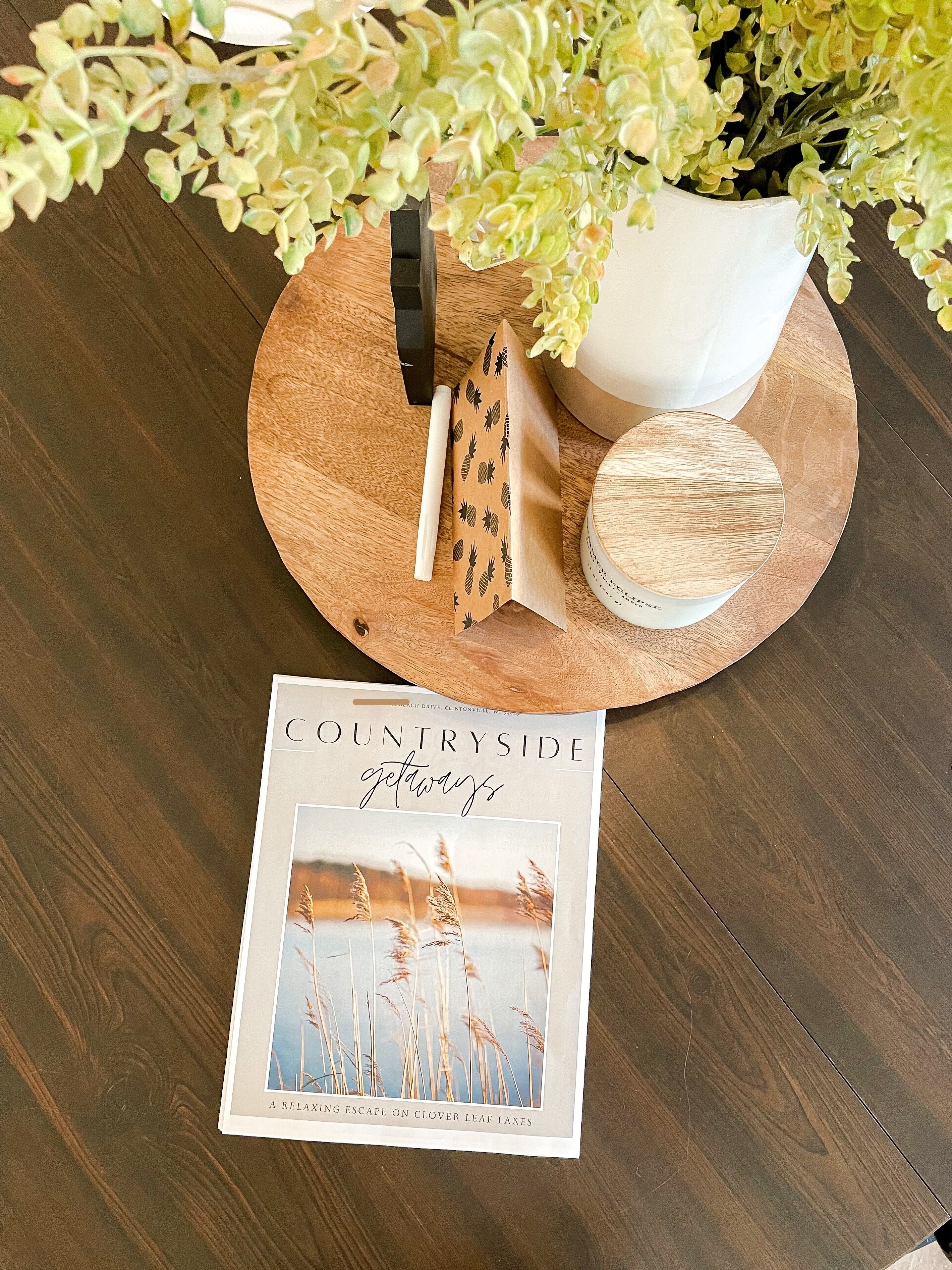 A wooden table with a brochure titled "COUNTRYSIDE getaways," a white vase with green foliage, and a wooden bowl.