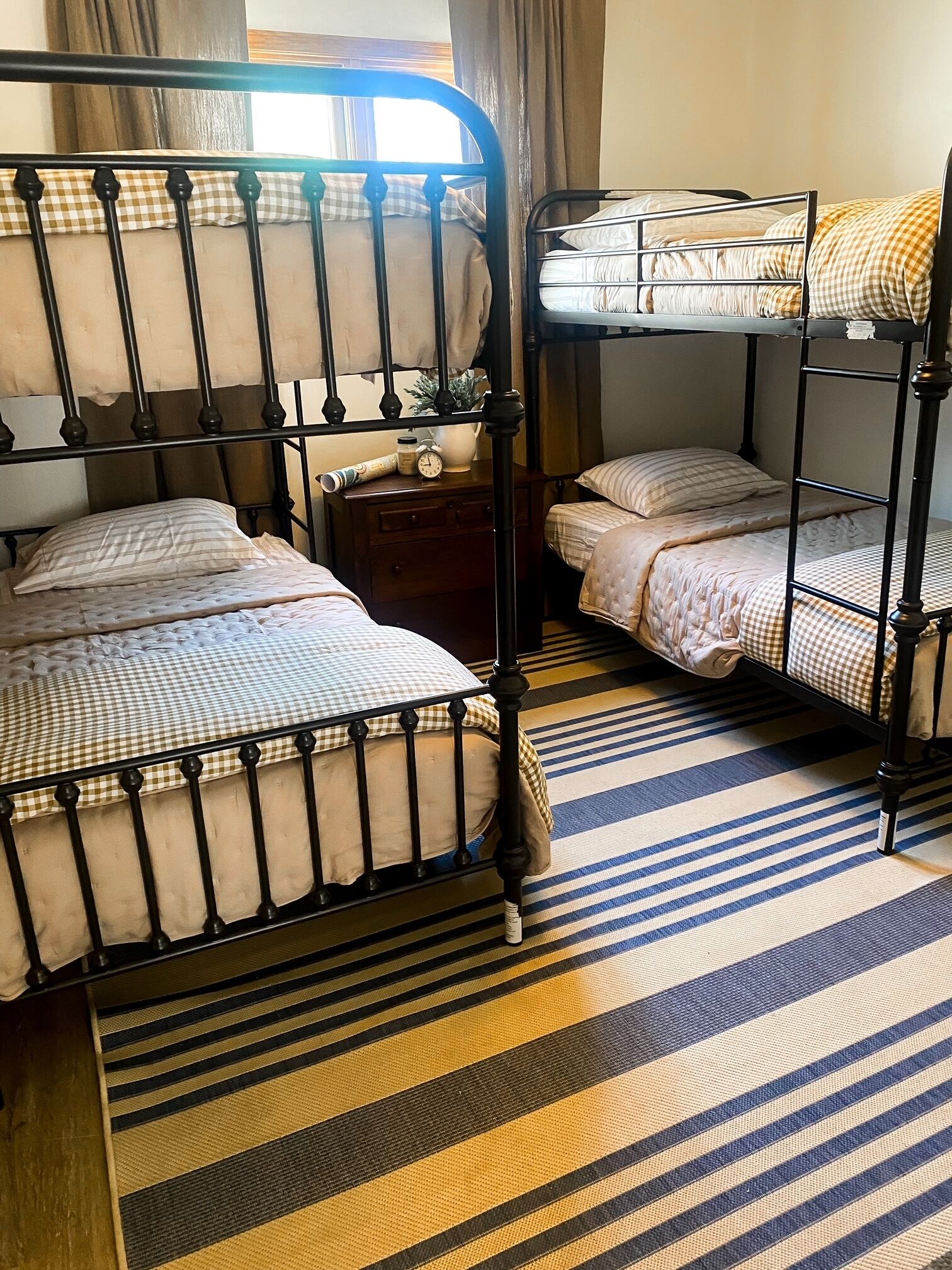 Une chambre avec un lit superposé, une couvre-lit à carreaux, une petite table de chevet avec une horloge et un tapis rayé.