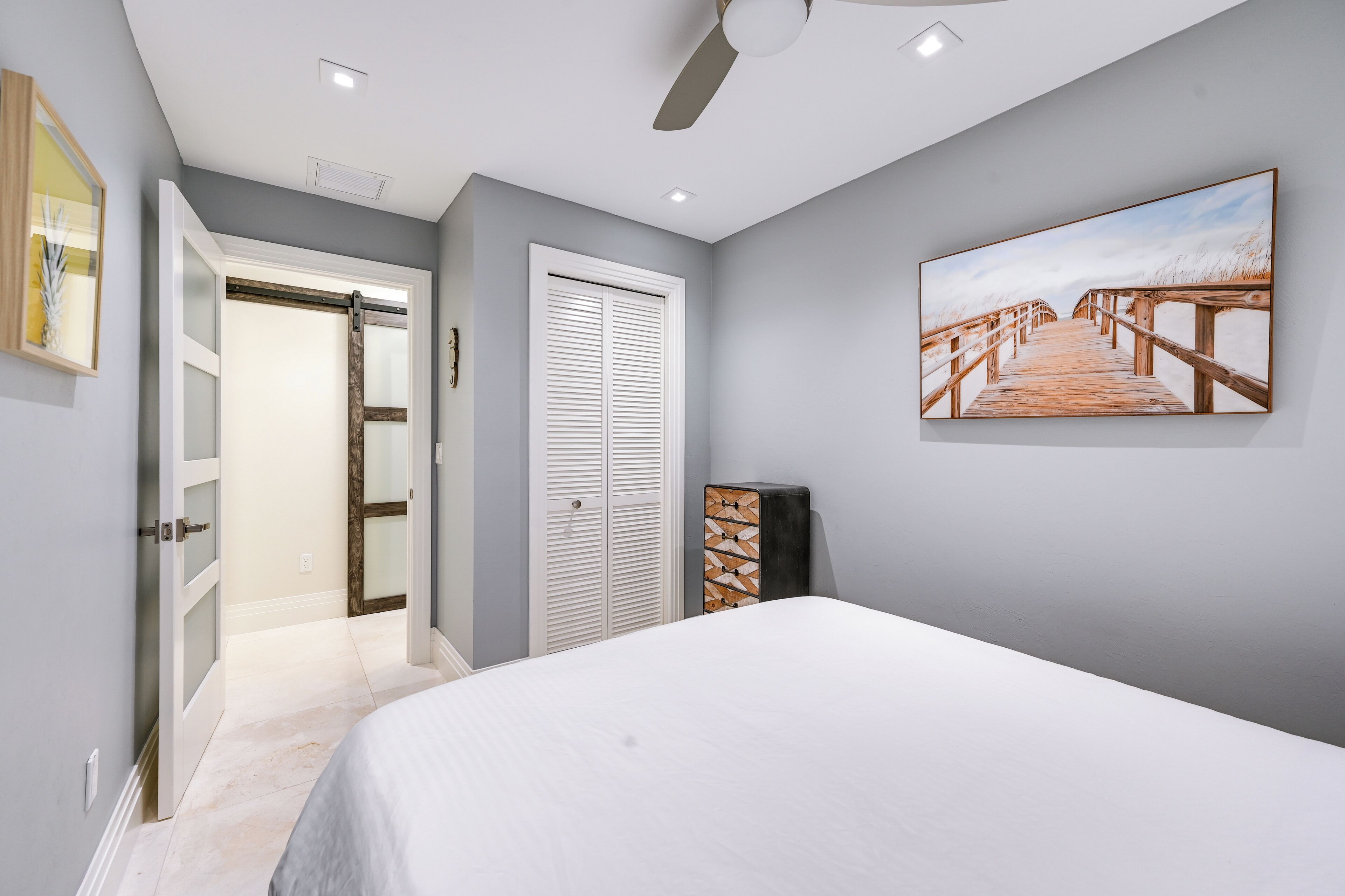 A bedroom with a bed, a ceiling fan, a painting of a beach boardwalk, and a closet with sliding doors.
