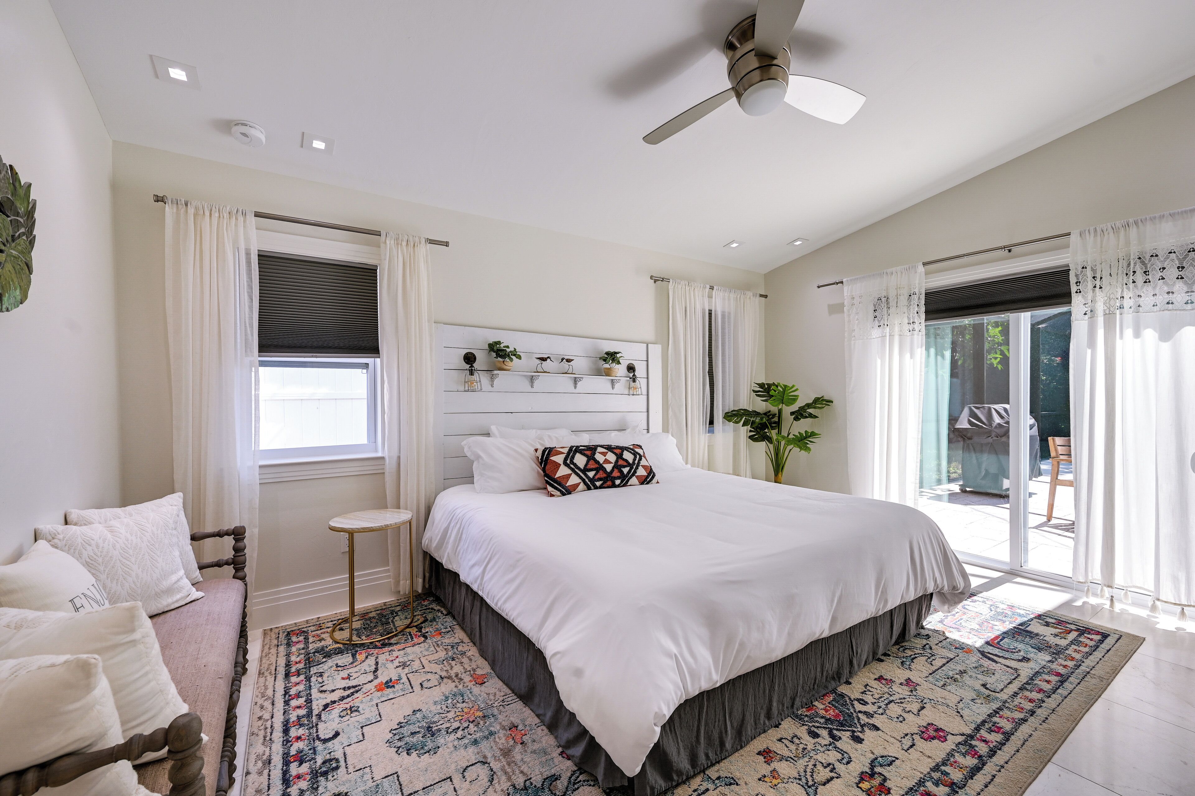 A bedroom with a large bed, a small bedside table, a ceiling fan, and a window with curtains.