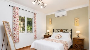 Une chambre à coucher comprenant un lit, deux tables de chevet, une fenêtre avec des rideaux, un climatiseur et des œuvres d’art accrochées aux murs.
