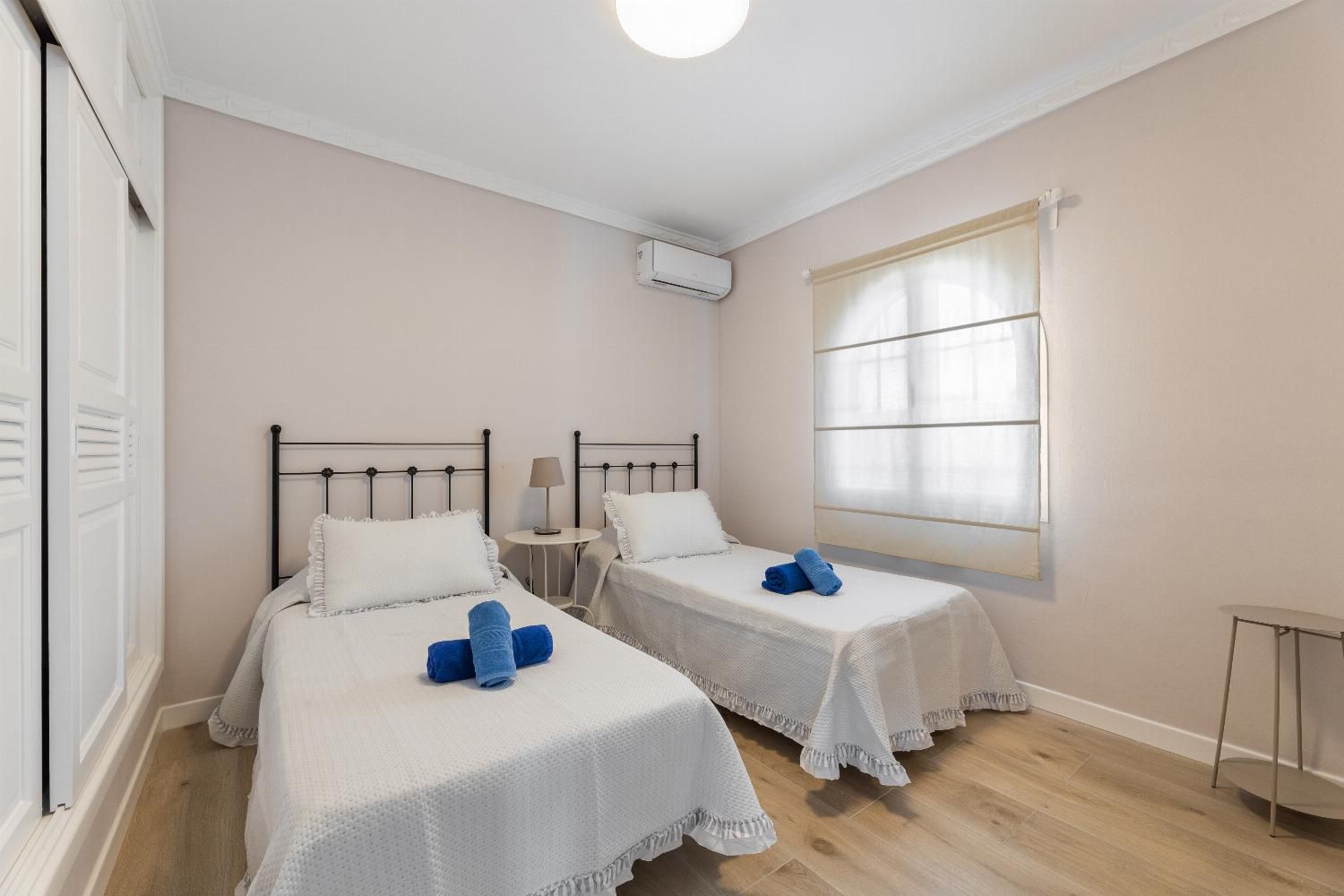 Ein Zimmer mit zwei Betten, einem kleinen Tisch, einem Fenster mit Vorhängen und einer Klimaanlage.