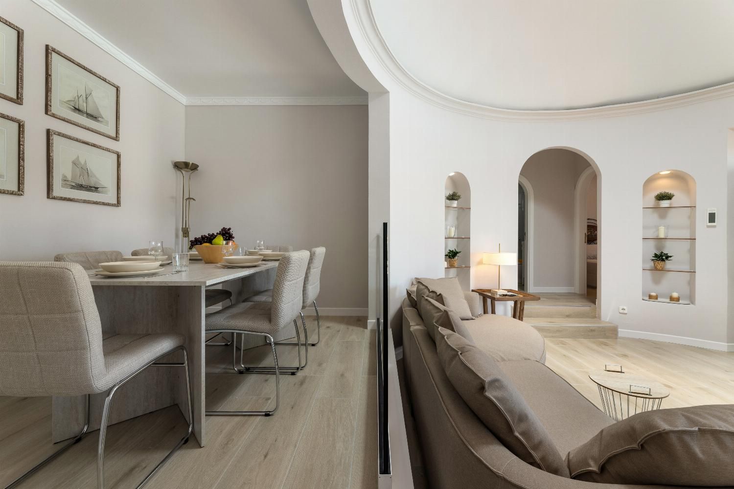 A modern dining area with a long table, chairs, and a sofa. There are framed pictures on the wall and a lamp on the table.