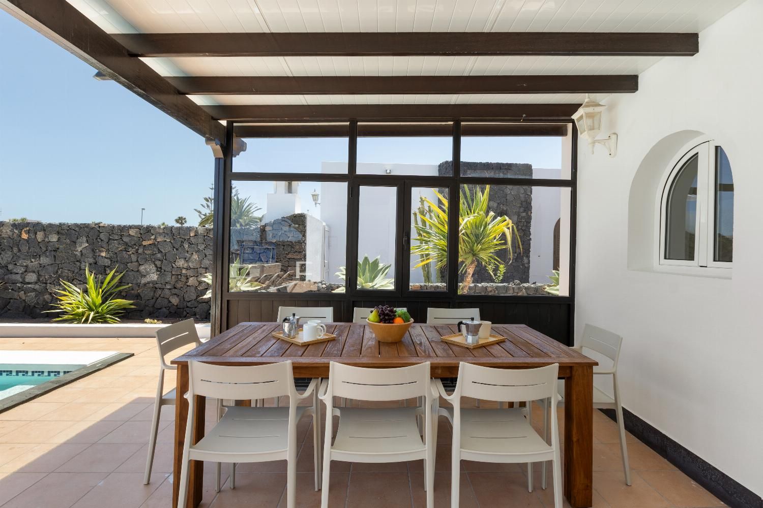 A dining area with a wooden table and white chairs, a stone wall, and a view of a pool and greenery.