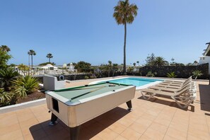 Mesa de bilhar, piscina, cadeiras de estar e céu claro.