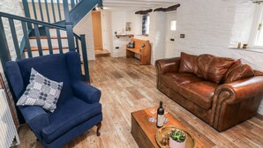 A living room with a blue armchair, a brown leather sofa, a wooden coffee table with a plant, and a staircase with blue railings. - Pensarngerrig (Llandysul)