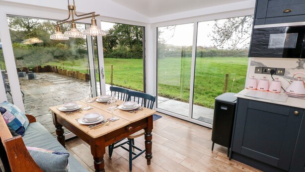 A kitchen with a dining table set for four, a built-in oven, and a view of a green garden through large sliding glass doors. - Pensarngerrig (Llandysul)