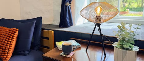 Een gezellige kamer met een houten bureau, een blauwe bank met oranje kussen, een plant in pot en een tafellamp.
