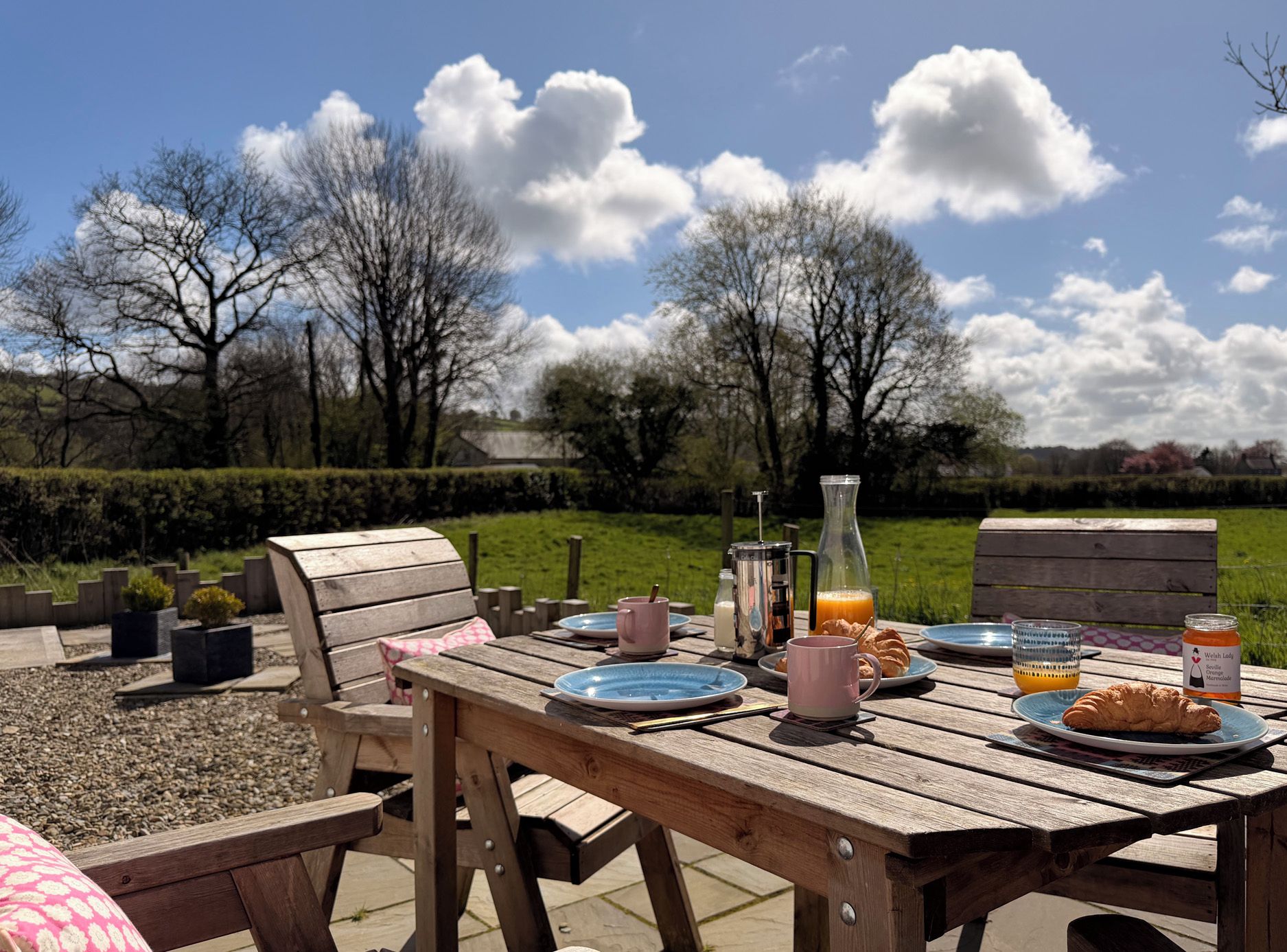 Un área de comedor al aire libre con una mesa de madera dispuesta para el desayuno, incluyendo platos, tazas y una botella de jugo de naranja.