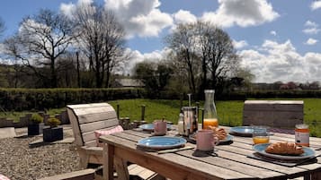 Un área de comedor al aire libre con una mesa de madera dispuesta para el desayuno, incluyendo platos, tazas y una botella de jugo de naranja.