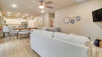 A modern living room with a sofa, dining table, and chairs. There is a ceiling fan, recessed lighting, and a kitchen in the background.
