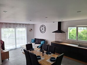 Moderni keittiö, jossa on suuri ikkuna, ruokailutila puupöydällä ja -tuoleilla sekä musta seinäkello.