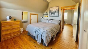 Een slaapkamer met een houten dressoir, een bed met een grijze deken, een hertenschilderij aan de muur en een plafondventilator.