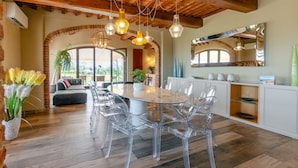 A spacious dining area with a glass table and chairs, a wooden ceiling, and a large window offering a view of the outdoors. - Villa Tosca (Peccioli)