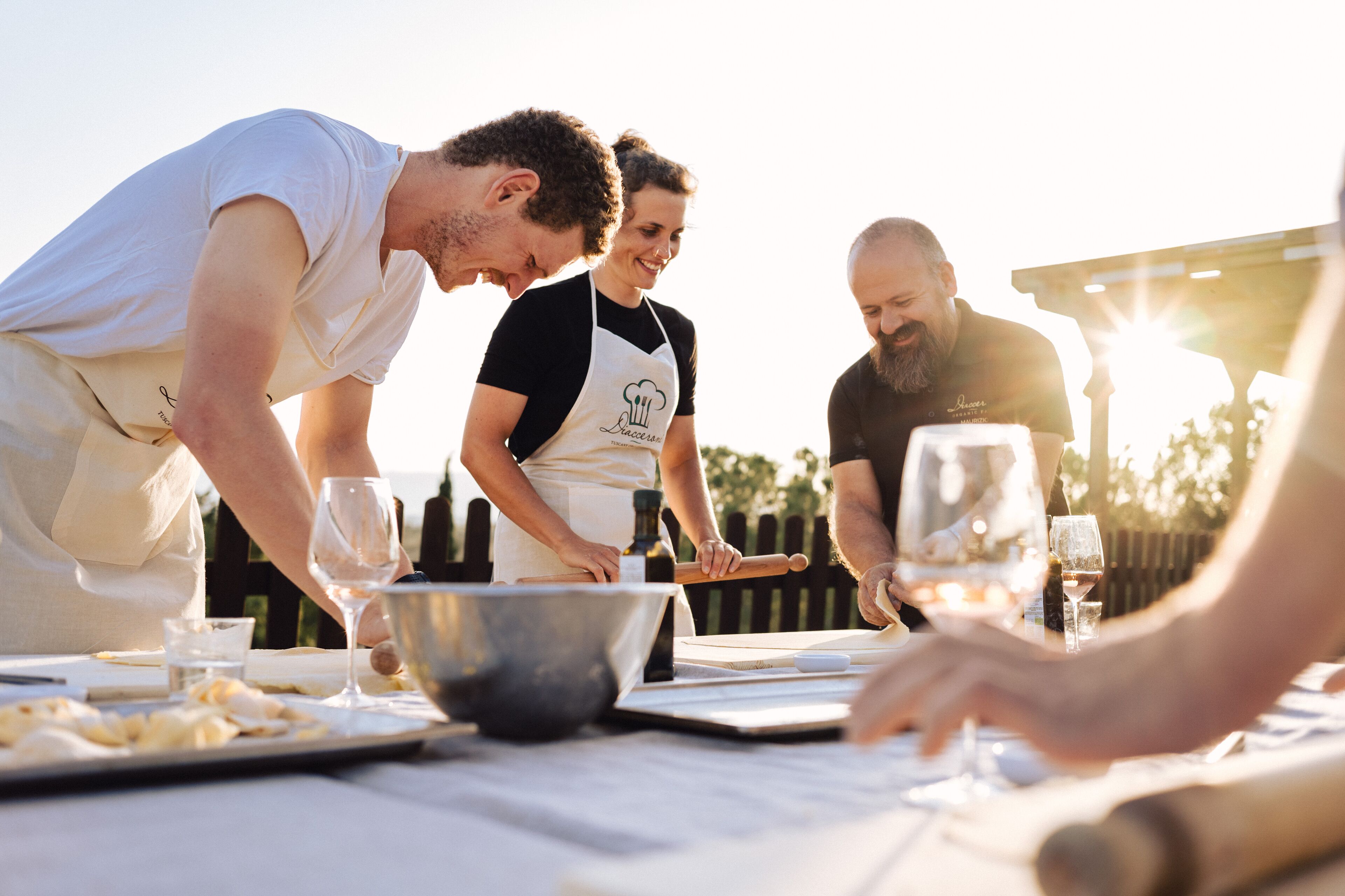 A group of people cooking outdoors with a table set for a meal, including wine glasses and a rolling pin.