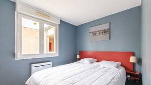 A bedroom with a bed, a window, a radiator, a wall-mounted picture, and a nightstand with a lamp. - Le Mayflower - One Bedroom Apartment, Sleeps 4 (Le Grau-du-Roi)