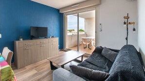 A modern living room with a sofa, a TV on a wooden cabinet, a dining area with a table and chairs, and a balcony with a view. - Le Mayflower - One Bedroom Apartment, Sleeps 4 (Le Grau-du-Roi)