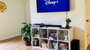 A living room with a flat-screen TV mounted on the wall displaying the Disney+ logo, a white entertainment unit with various items, and a wooden floor. - Cozy DFW Duplex, central location, kids, pets (North Richland Hills)
