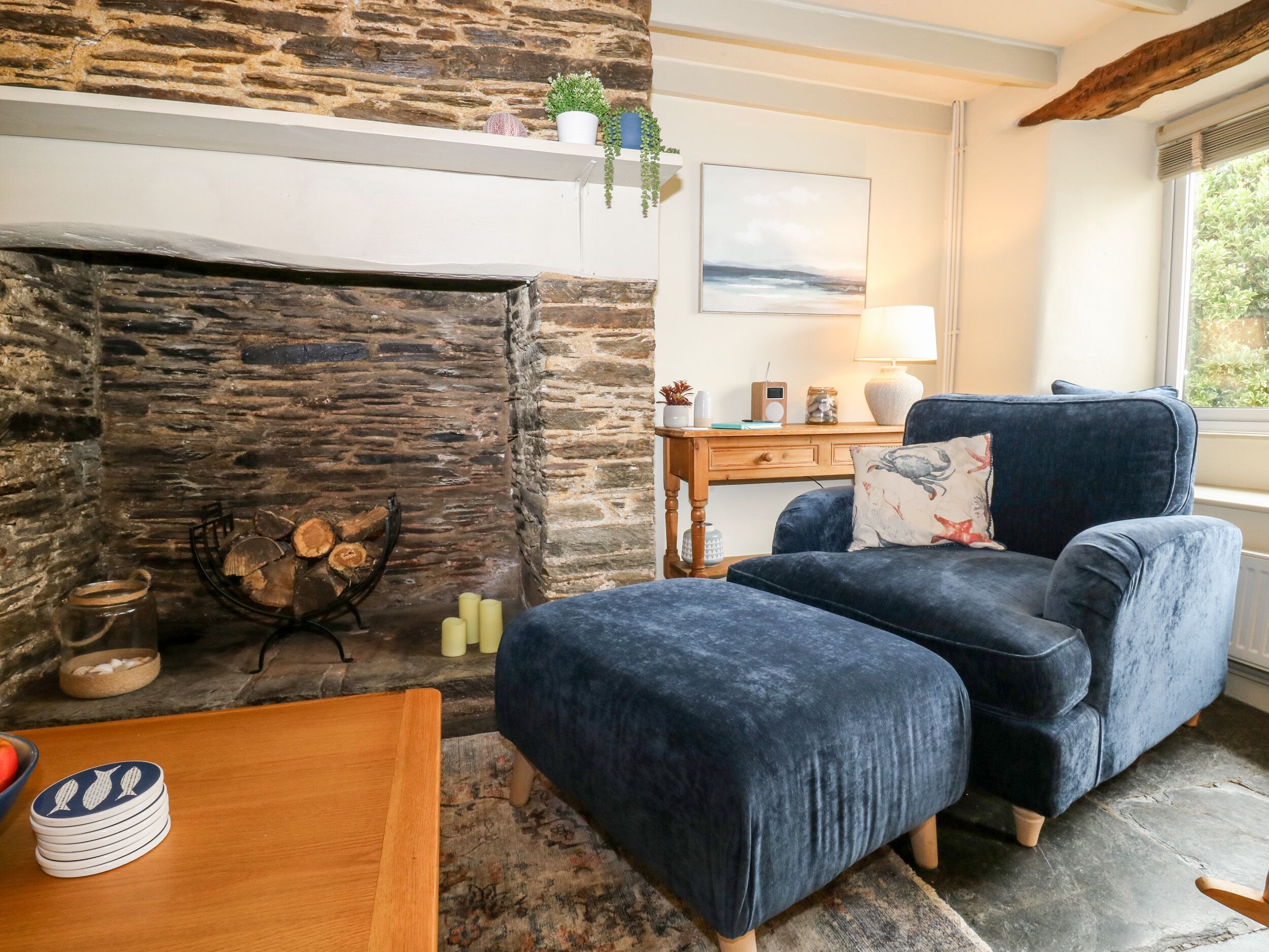 Een gezellige woonkamer met een stenen openhaard, een blauwe fauteuil, een houten salontafel en uitzicht op groen.