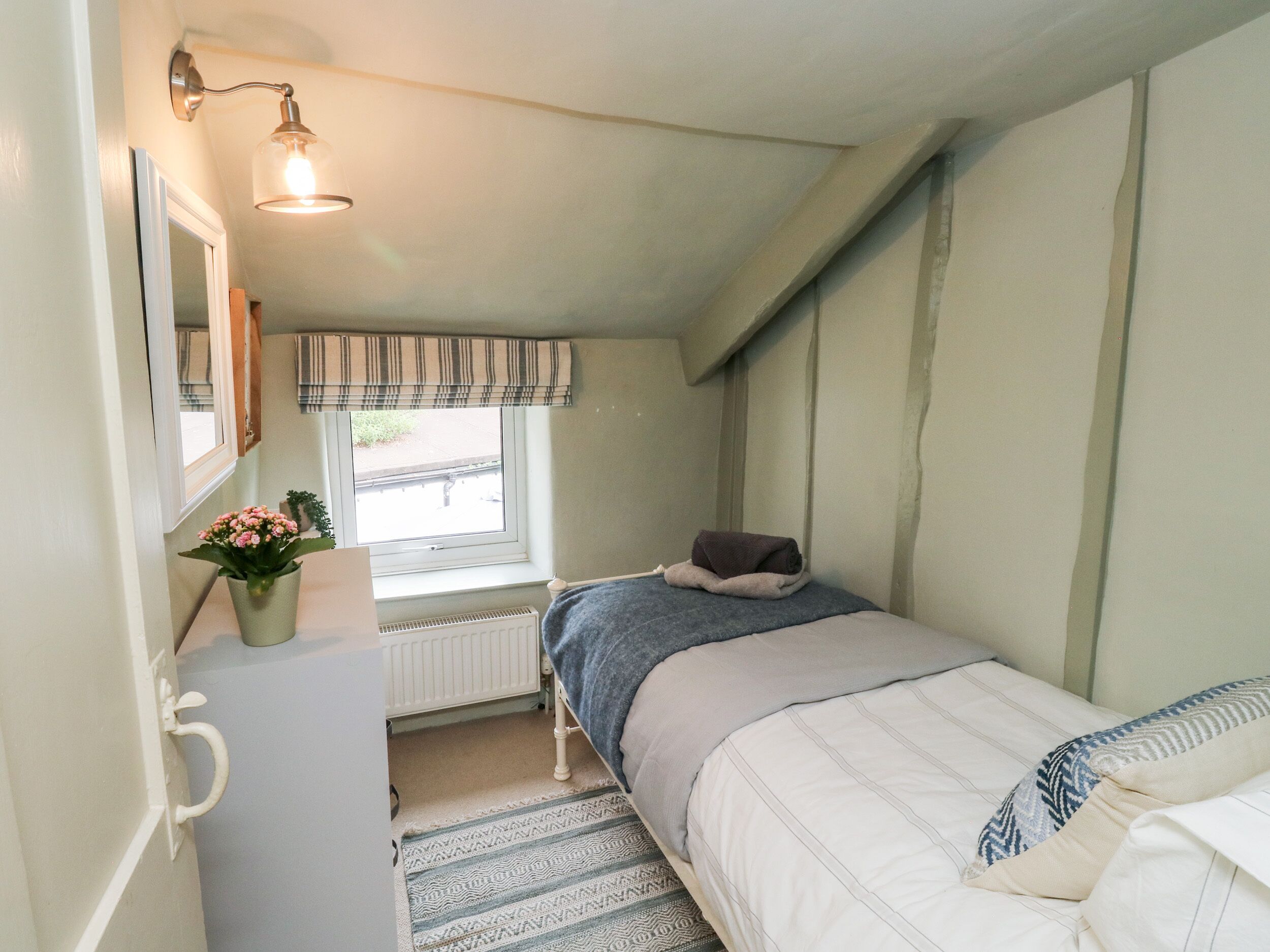 A bedroom with a bed, a radiator, a window with curtains, a bedside table with a plant, and a wall-mounted light fixture.