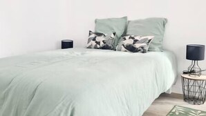 Un lit avec une couvre-lit verte et deux oreillers, une petite table de chevet avec une lampe et un haut-parleur fixé au mur.