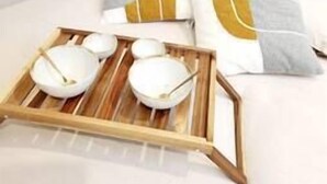 Ein Bett mit weißer Bettwäsche, zwei Kissen mit einem gelb-grauen Muster und ein Holztablett mit Schüsseln und Löffeln.