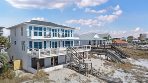 A two-story beachfront house with a balcony, a deck, and a staircase leading to the beach.