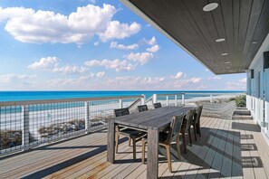 Een houten terras met een tafel en stoelen, met uitzicht op een strand en zee.