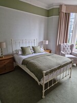 Un dormitorio ordenado con cama, mesitas de noche, una silla y una ventana con cortinas.