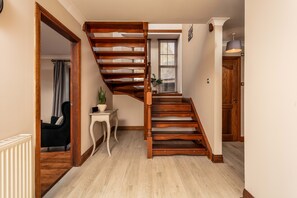 A wooden staircase with a white railing, a small table with a plant, and a chair in a hallway with light-colored flooring. - Cromwell Corner - Donnini Apartments  (Ayr)