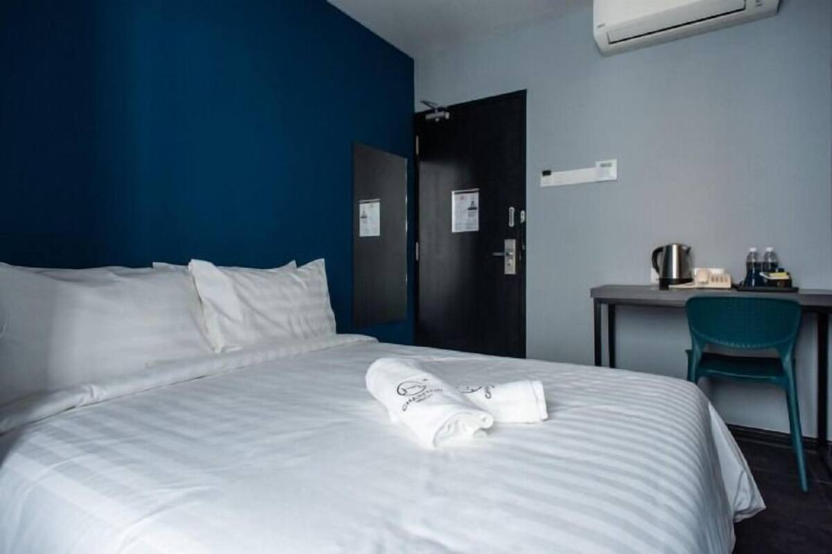A hotel room with a bed, a desk, and a chair. There is a door and an air conditioning unit on the wall.