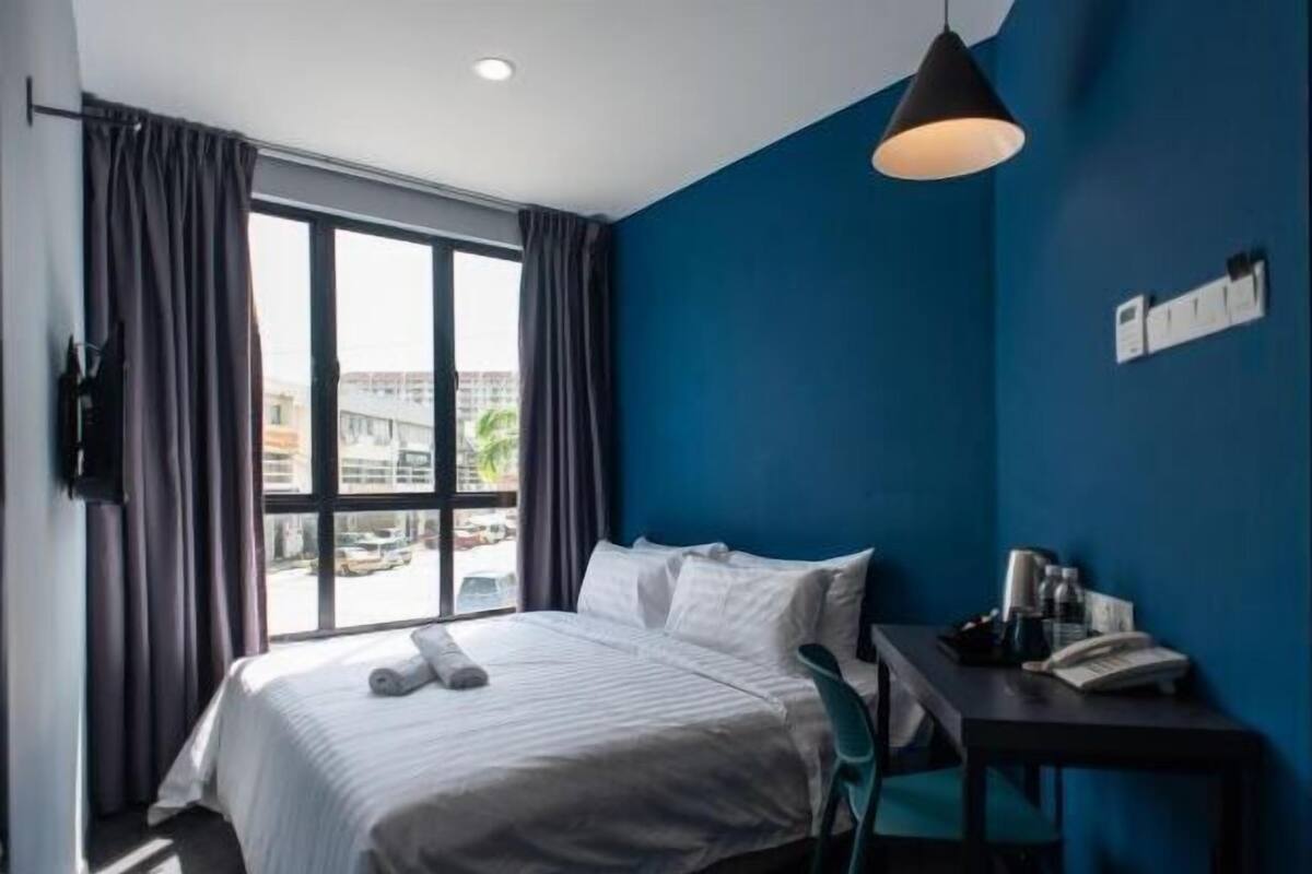 A hotel room with a bed, a desk with a chair, a window with curtains, and a hanging light fixture.