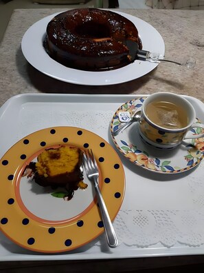 A cake on a white plate with a fork, a cup of tea on a saucer, and a decorative plate with a fruit pattern. - The Spot - Arraial do Cabo (Arraial do Cabo)