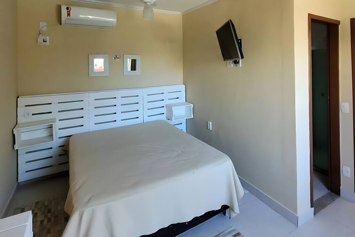 A bedroom with a bed, a ceiling fan, an air conditioning unit, a wall-mounted TV, and a door.