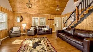 Un vaste salon aux murs et au plafond en bois, avec un canapé en cuir marron, deux fauteuils, une table basse et un grand tapis orné d’un motif d’ours.