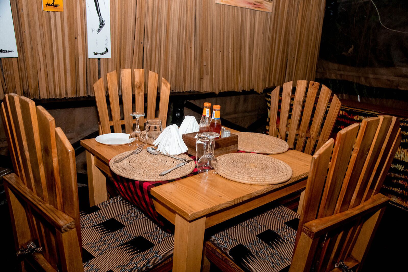 A wooden dining table set with plates, cutlery, and bottles, surrounded by wooden chairs with patterned cushions.