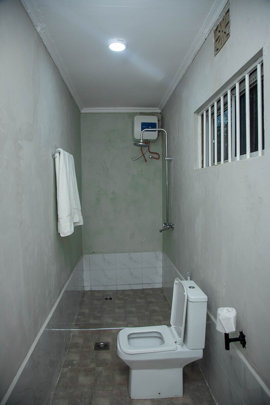 A small, narrow bathroom with a toilet, a shower, and a window with bars.