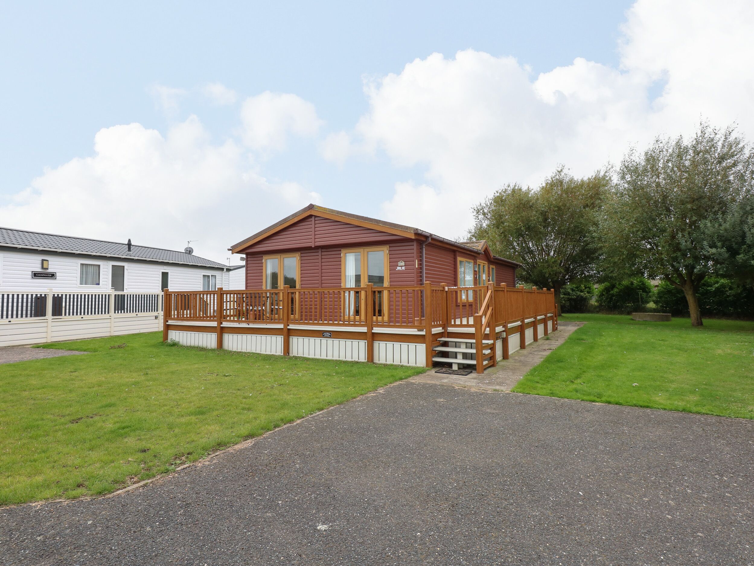 A single-story mobile home with a wooden deck and a small porch, surrounded by a grassy area and a paved path.