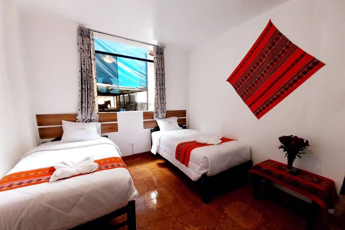 A hotel room with two beds, a window with curtains, a wall hanging, and a small table with a vase.