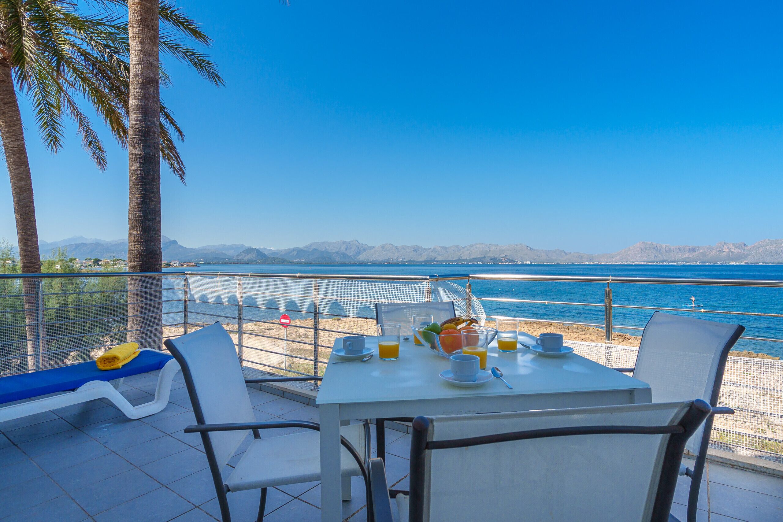 A dining table with chairs and a bench, overlooking a beach and mountains.
