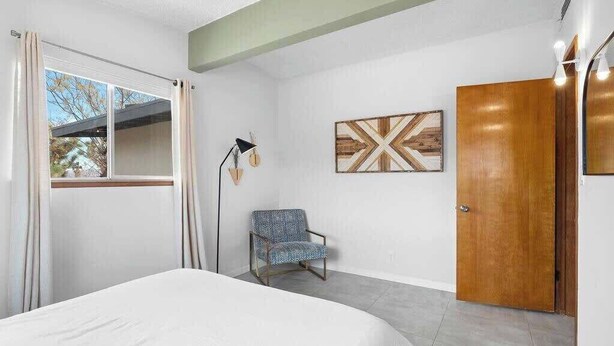 A bedroom with a bed, a chair, a wooden door, a window with curtains, and a wall-mounted lamp. - Moonrise Ranch|Yoga Sun Room|Cowboy Pool (Joshua Tree)