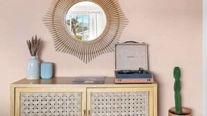 A room with a circular mirror, a small cabinet with woven doors, a cactus plant, and a suitcase on the cabinet. - Moonrise Ranch|Yoga Sun Room|Cowboy Pool (Joshua Tree)
