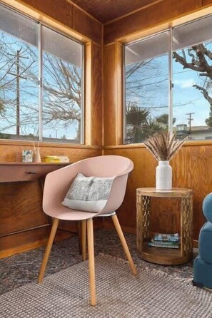 A corner room with a wooden wall, a chair with a cushion, a small wooden side table, and a window with a view of trees and a building.