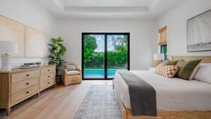 Une chambre moderne avec un grand lit, une commode, une plante et une vue sur une piscine, visible à travers une porte vitrée.