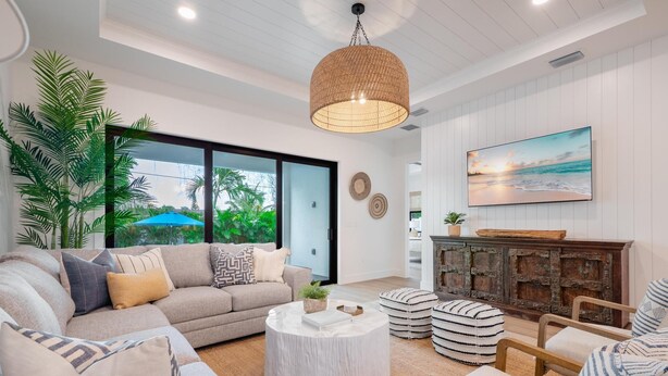 Un salon moderne avec un grand canapé, une table basse ronde et des poufs à rayures. Une grande fenêtre offre une vue sur la verdure à l’extérieur.