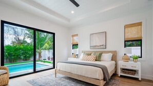Une chambre moderne avec un grand lit, une table de chevet, une fenêtre donnant sur une piscine et un espace verdoyant, ainsi qu’un ventilateur de plafond.
