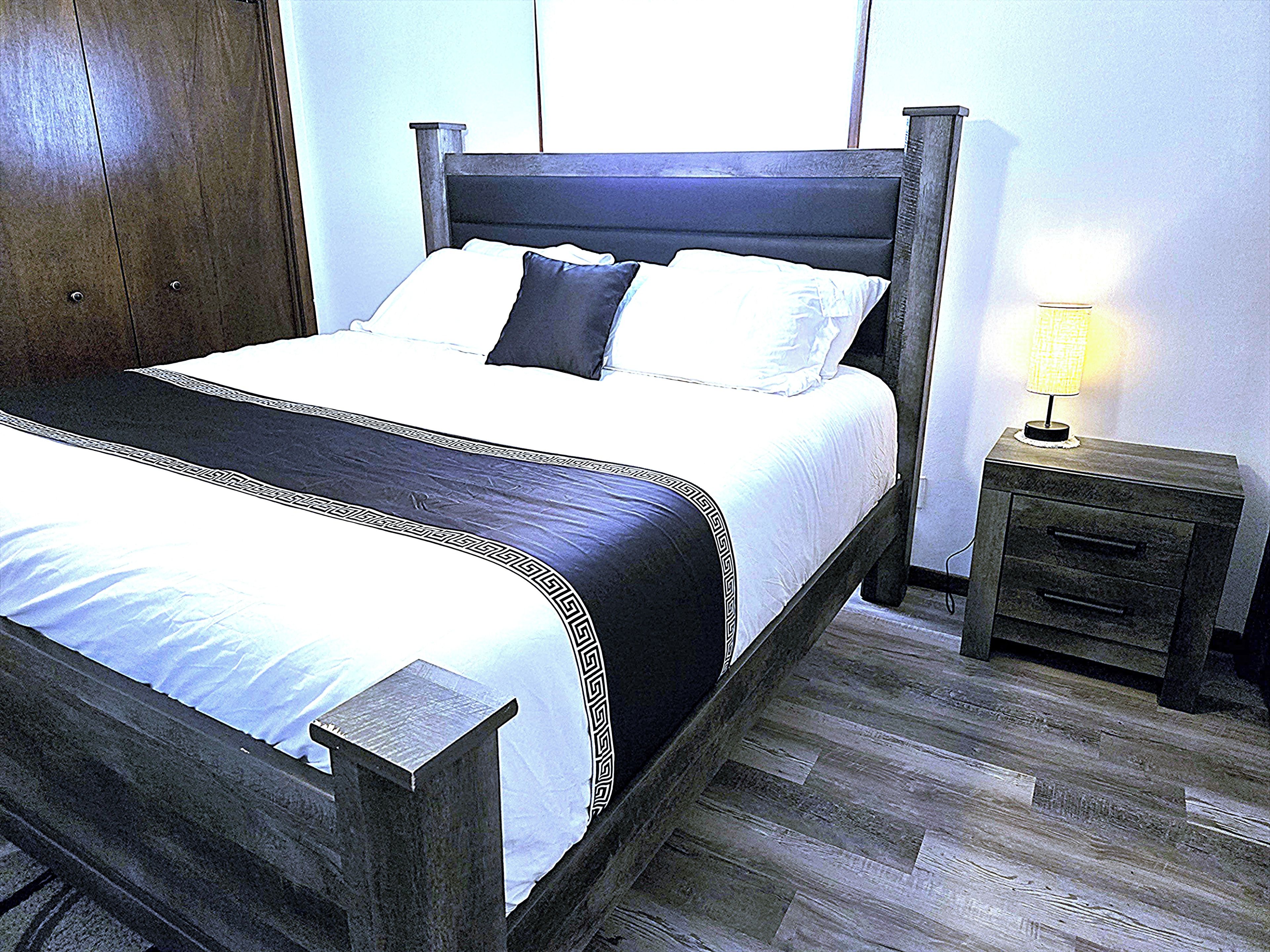 Een bed met een donker hoofdbord, wit beddengoed en een decoratieve rand. Er is een nachtkastje met een lamp en een lade. De kamer heeft houten vloeren en een houten deur.